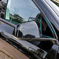 Close-up of a glossy black car side mirror and window trim with a thin blue accent line, reflecting an outdoor city scene.